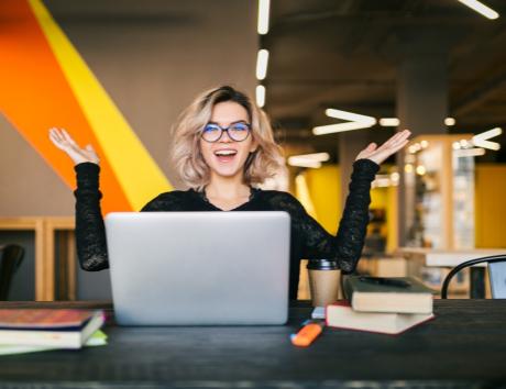 Das Bild zeigt eine fröhliche Frau mit Laptop; außerdem zu sehen: einige Bücher und Dokumente, ein Marker und ein Kaffeebecher