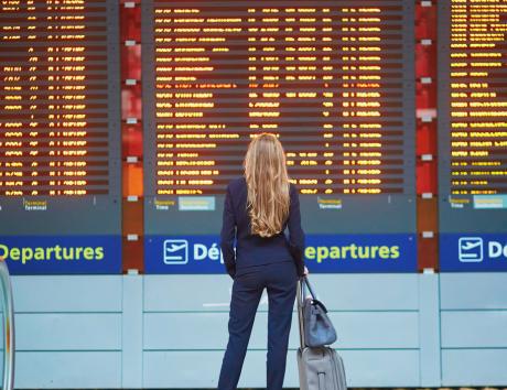 Une femme est debout dans un aéroport devant trois tableaux d'affichage montrant les départs. Elle porte un tailleur et a deux sacs symbolisant un déménagement dans une autre région pour raisons professionnelles.