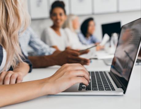 Grupa pracownikach siedząca w jednym rzędzie przy komputerach, na pierwszym planie ucięta postać kobiety, której ręka pisze na klawiaturze laptopa.