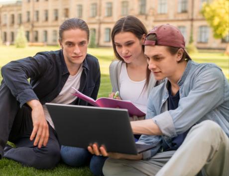 Das Bild zeigt eine Gruppe von Studenten, die vor einem Gebäude auf dem Rasen sitzen und sich auf einem Laptop etwas ansehen.