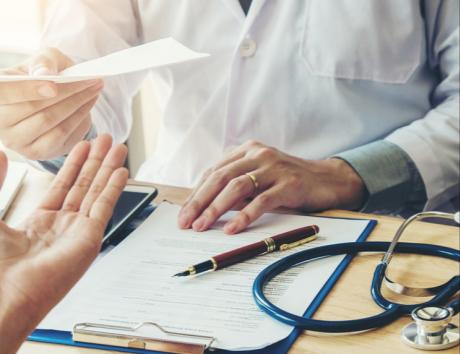 Das Bild zeigt eine Arztkonsultation, man sieht die Hände des Arztes in weißem Arztkittel und die Hände einer Patientin. Außerdem einem Diagnosebericht mit Kugelschreiber sowie Stethoskop auf dem Tisch zwischen den beiden.